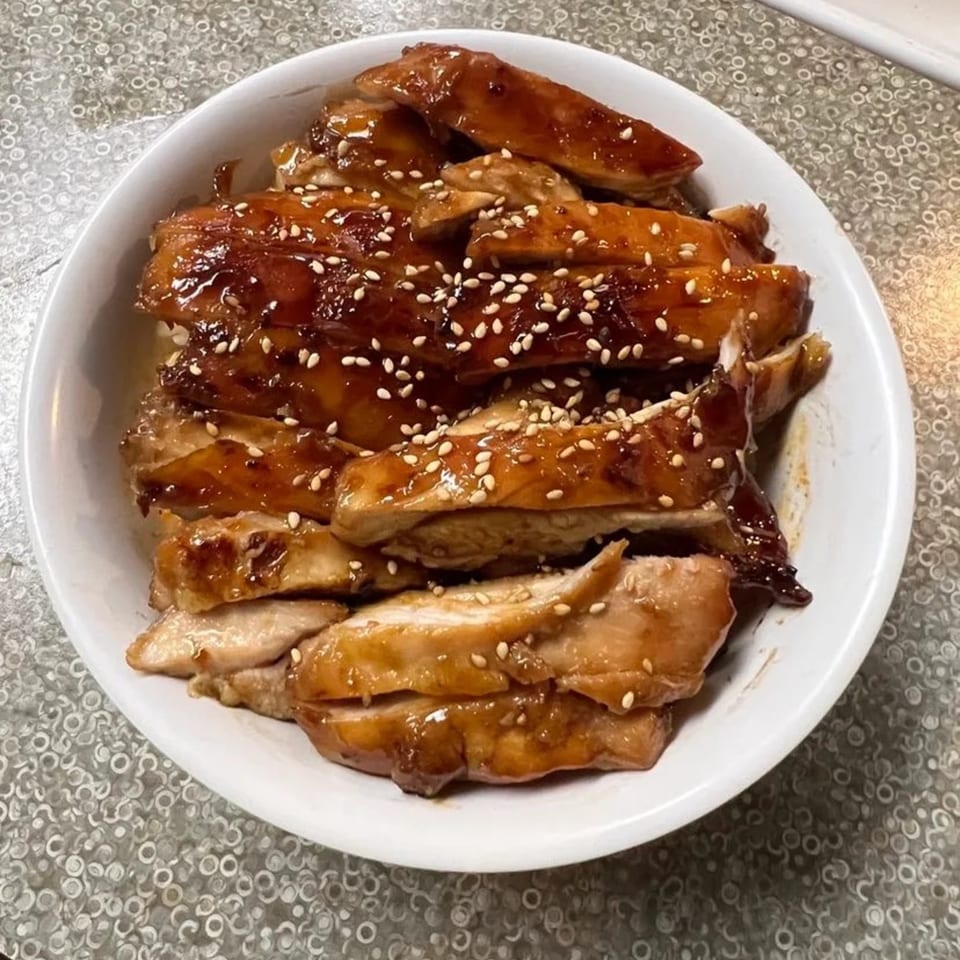 Teriyaki chicken slices, glistening with sauce and sprinkled with sesame seeds, in a white bowl.